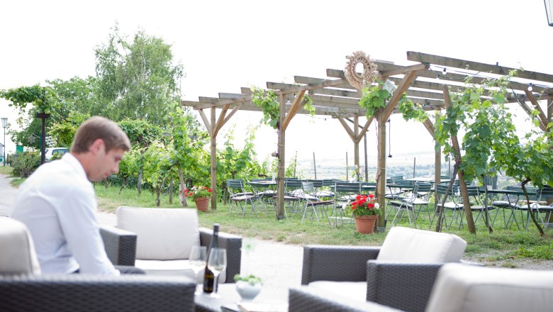 Gastgarten, © Bernd Baumgartner Ein Mann sitzt in einem Gastgarten mit Weinreben und leeren Tischen im Hintergrund.