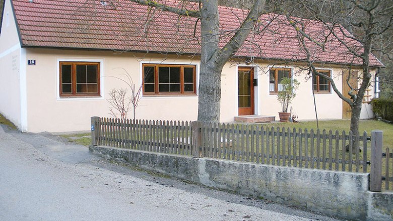 Ferienhaus im Loistal, © Ursin Haus Ein einstöckiges Ferienhaus mit rotem Ziegeldach und hellem Anstrich, umgeben von einem Holzzaun und Bäumen.