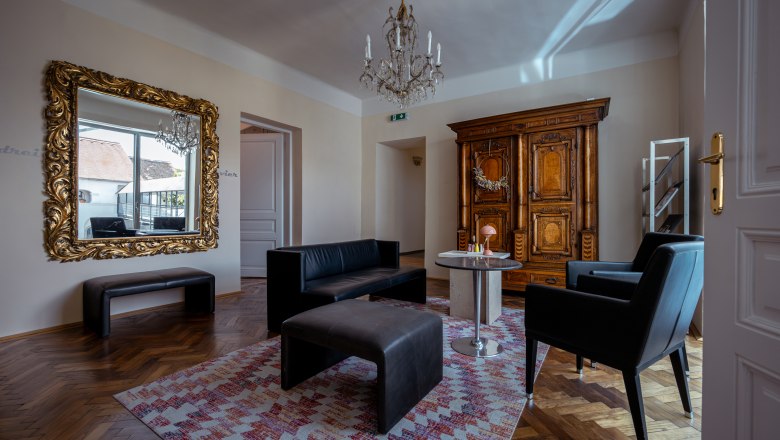 vierzigerhof, © Niederösterreich Werbung / Maximilian Pawlikowsky Elegantes Wohnzimmer mit antikem Schrank, großem Spiegel, schwarzen Ledermöbeln und einem Kronleuchter.