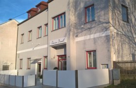 Pension Brandl, © Pension Brandl Außenansicht der Pension Brandl mit grauer Fassade und roten Fensterrahmen.