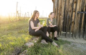 Weingut Grillmaier, © Weingut Grillmaier Zwei Frauen sitzen mit Weingläsern vor einer Holzhütte im Weinberg.