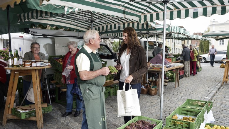 Wochenmarkt am Kornplatz, © POV, Robert Herbst Menschen auf einem Wochenmarkt mit Obst- und Gemüseständen unter gestreiften Markisen.