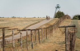 Eiserner-Vorhang-Cizov, © Waldviertel Tourismus Zwei Radfahrer auf einem Weg neben einem alten Grenzzaun und einem Wachturm.