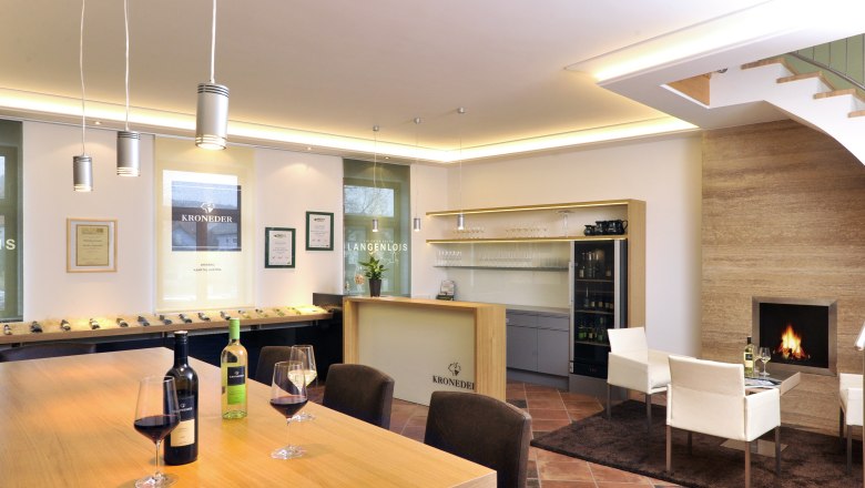Wein & Wohnen Kroneder, © Familie Kroneder Innenansicht eines modernen Weinverkostungsraums mit Holztisch, Weingläsern, Kamin und Regalen.