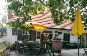 Gemütlicher Gastgarten mit gelbem Sonnenschirm und Holztischen vor einem Gebäude mit rotem Ziegeldach.