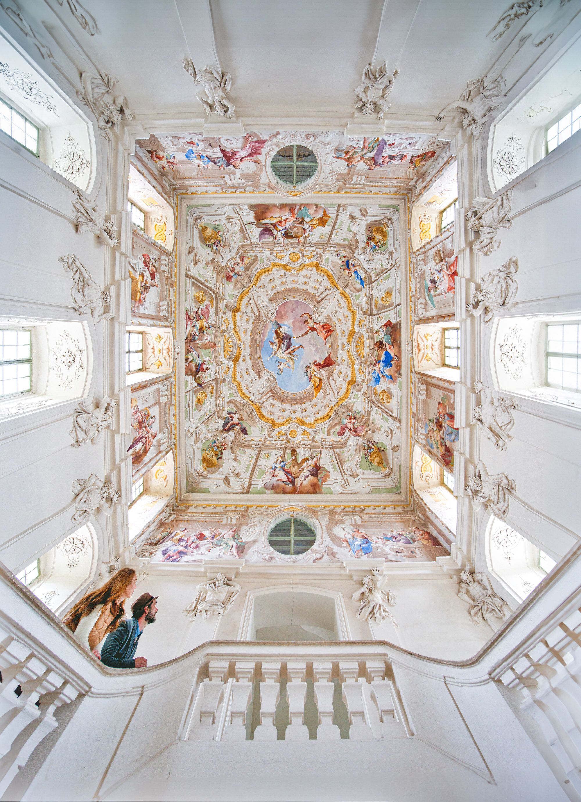 Zwei Personen betrachten die kunstvoll bemalte Decke eines barocken Treppenhauses im Stift Altenburg.