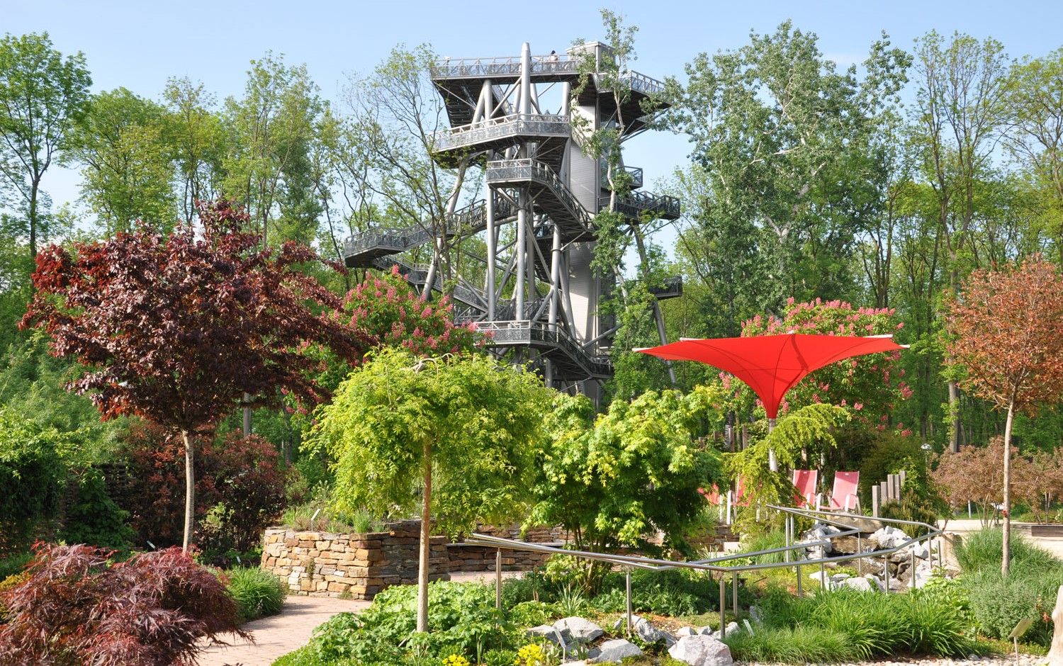 Ein Baumwipfelpfad mit einem hohen, modernen Turm aus Metall in einem grünen Waldgebiet.