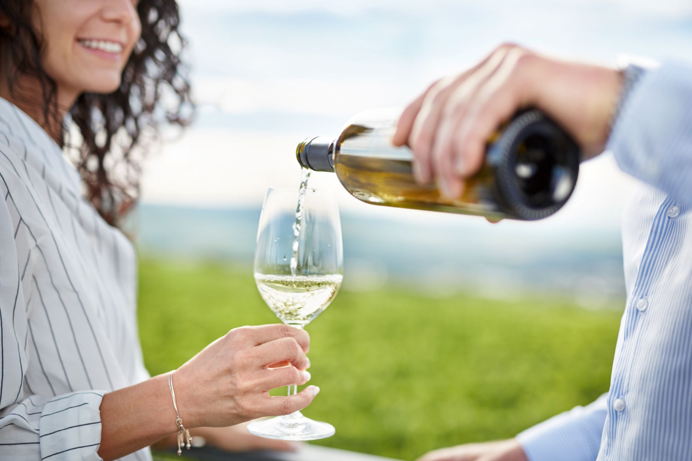 Eine Person schenkt Weißwein in ein Glas, das von einer anderen Person gehalten wird, im Hintergrund ein Weinberg.