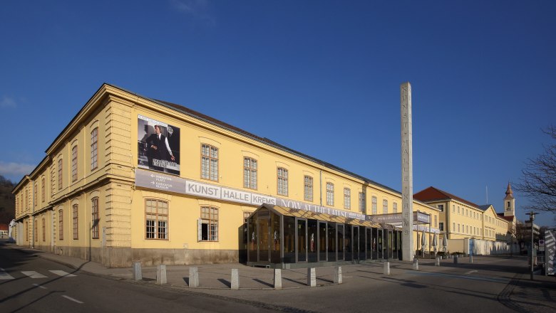 Außenansicht der Kunsthalle Krems, ein gelbes Gebäude mit großen Fenstern und einem Plakat an der Fassade.