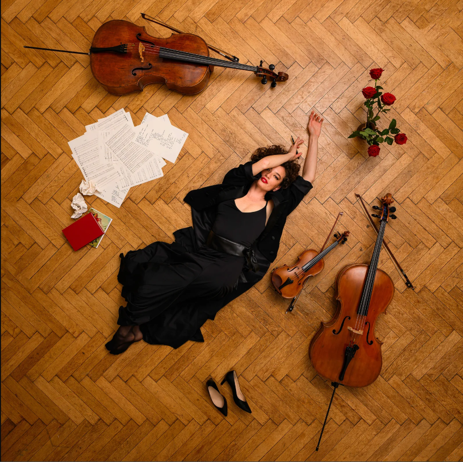 Eine Frau in schwarzem Gewand liegt am Boden, neben ihr eine Geige, ein Cello und Noten.