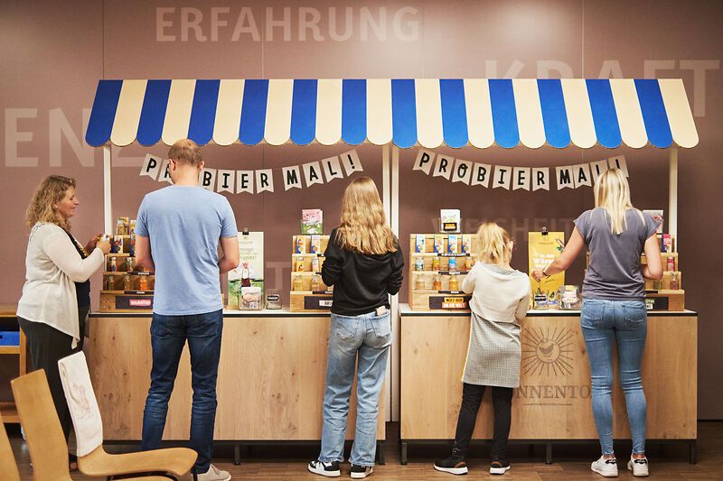 Menschen probieren Produkte an einem Stand mit blau-weißem Dach.