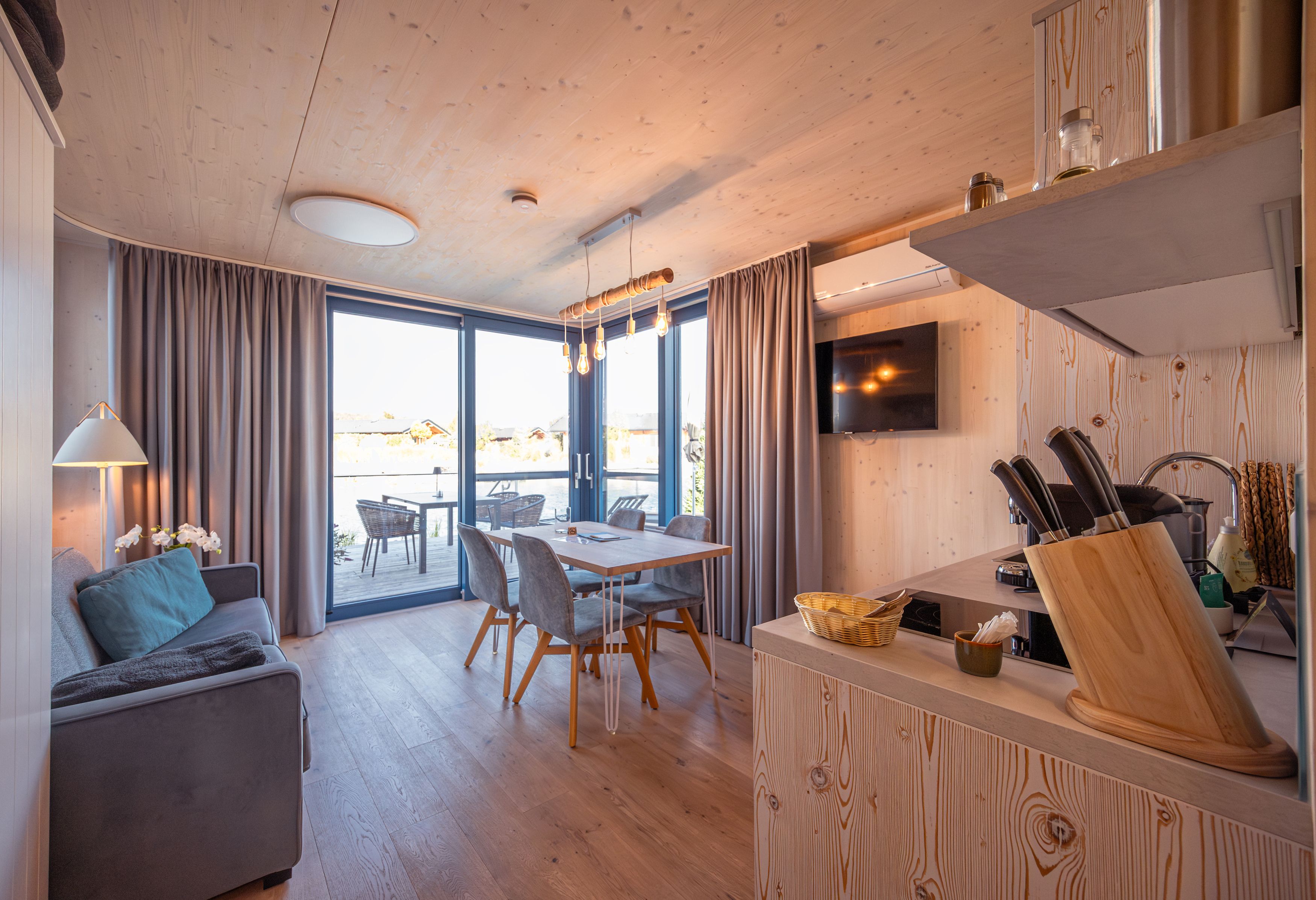 Innenansicht eines modernen Chalets mit Holzwänden, Esstisch, Sofa und großer Fensterfront mit Blick auf eine Terrasse.