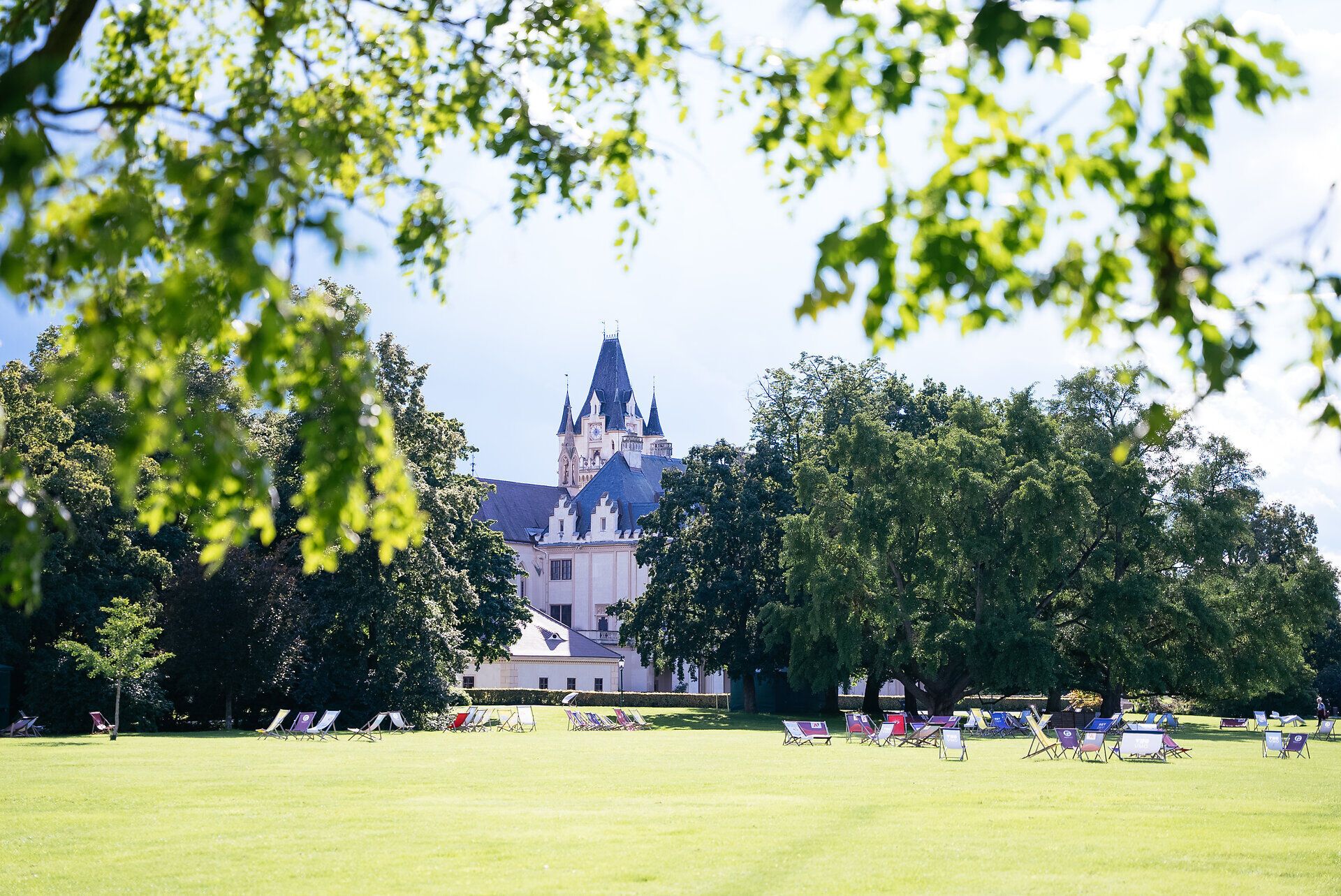 Die weitläufige Wiese lädt zum Entspannen ein, während majestätische Bäume Schatten spenden. Im Hintergrund erhebt sich das beeindruckende Schloss, das mit seiner romantischen Architektur eine zauberhafte Kulisse bietet. Hier erleben Besucher die perfekte Harmonie von Natur und Kultur.