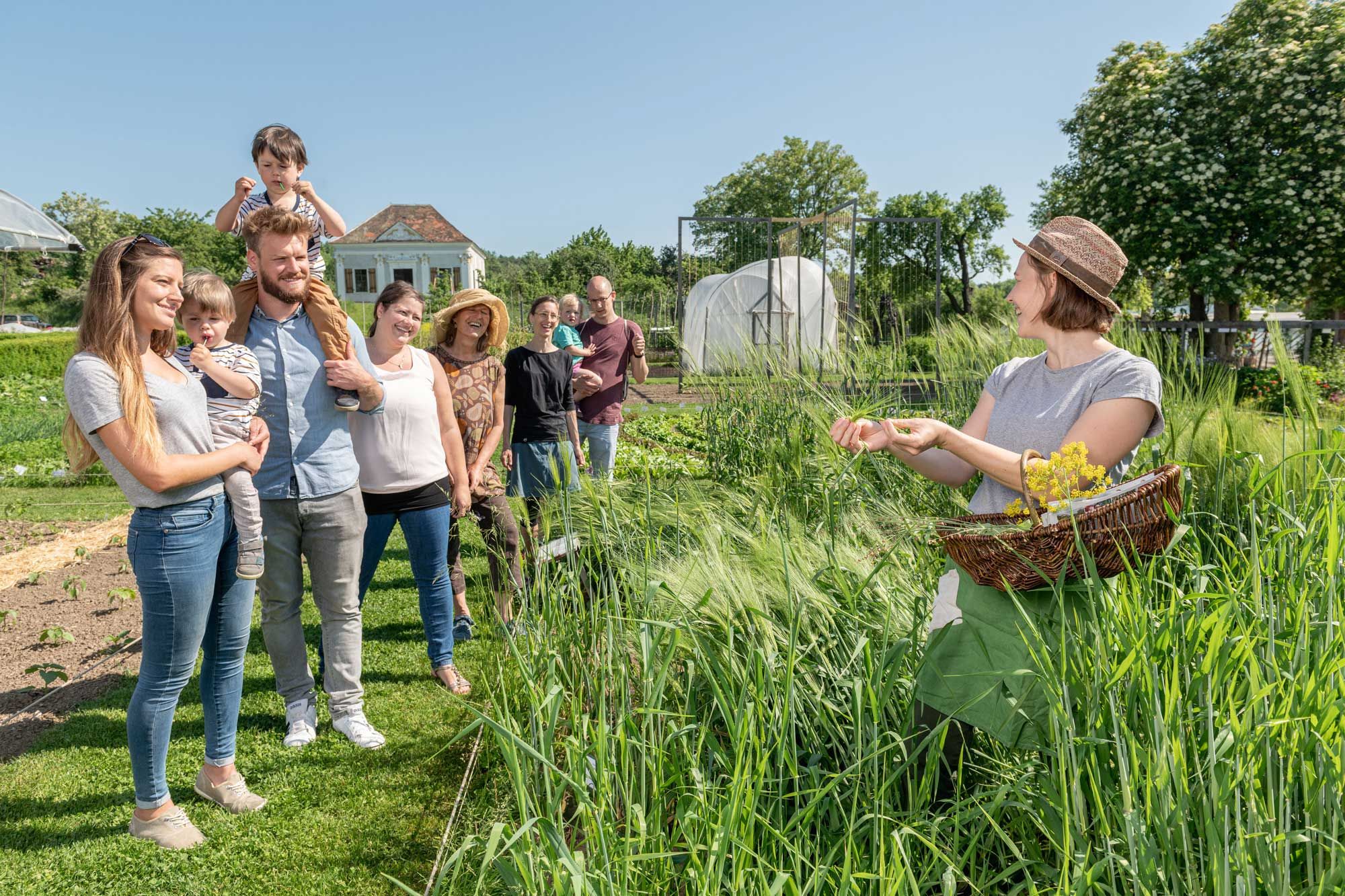 Eine Gruppe von Menschen steht in einem Garten, während eine Frau mit einem Korb Pflanzen zeigt.