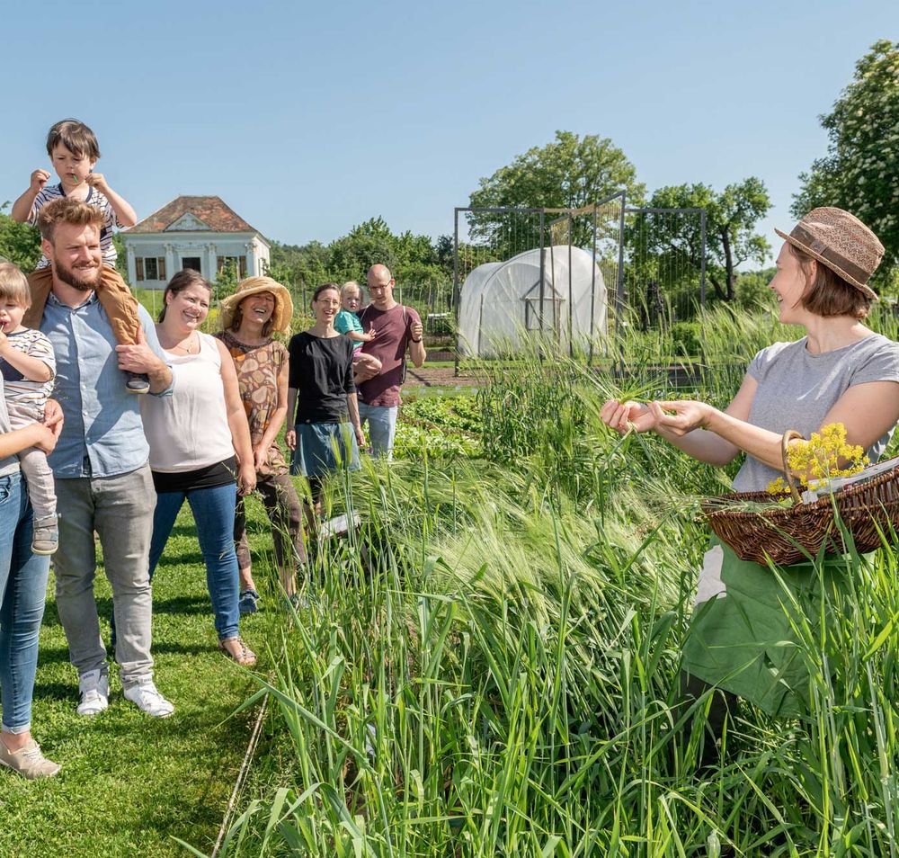 Eine Gruppe von Menschen steht in einem Garten, während eine Frau mit einem Korb Pflanzen zeigt.