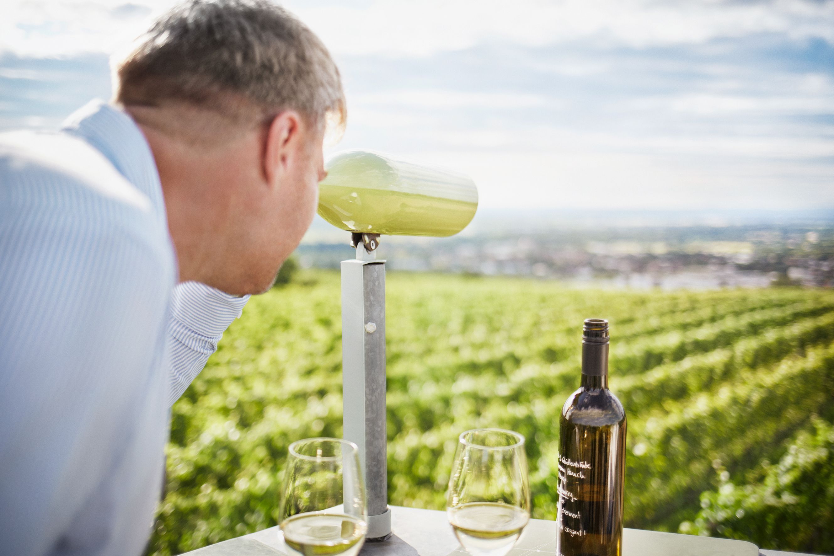 Ein Mann blickt durch ein Fernglas auf ein Weinfeld. Auf dem Tisch stehen eine Weinflasche und zwei Gläser.