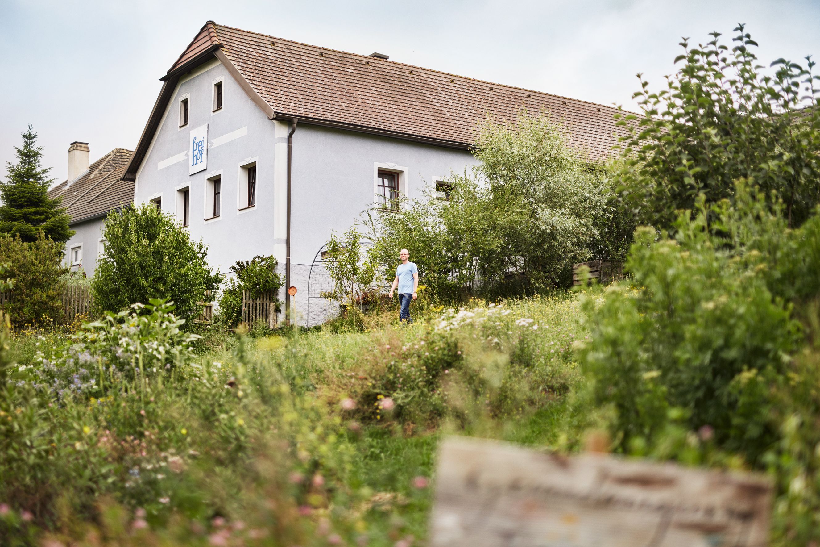 Ein Mann steht vor einem traditionellen österreichischen Haus, umgeben von üppigem Grün und einem Garten.