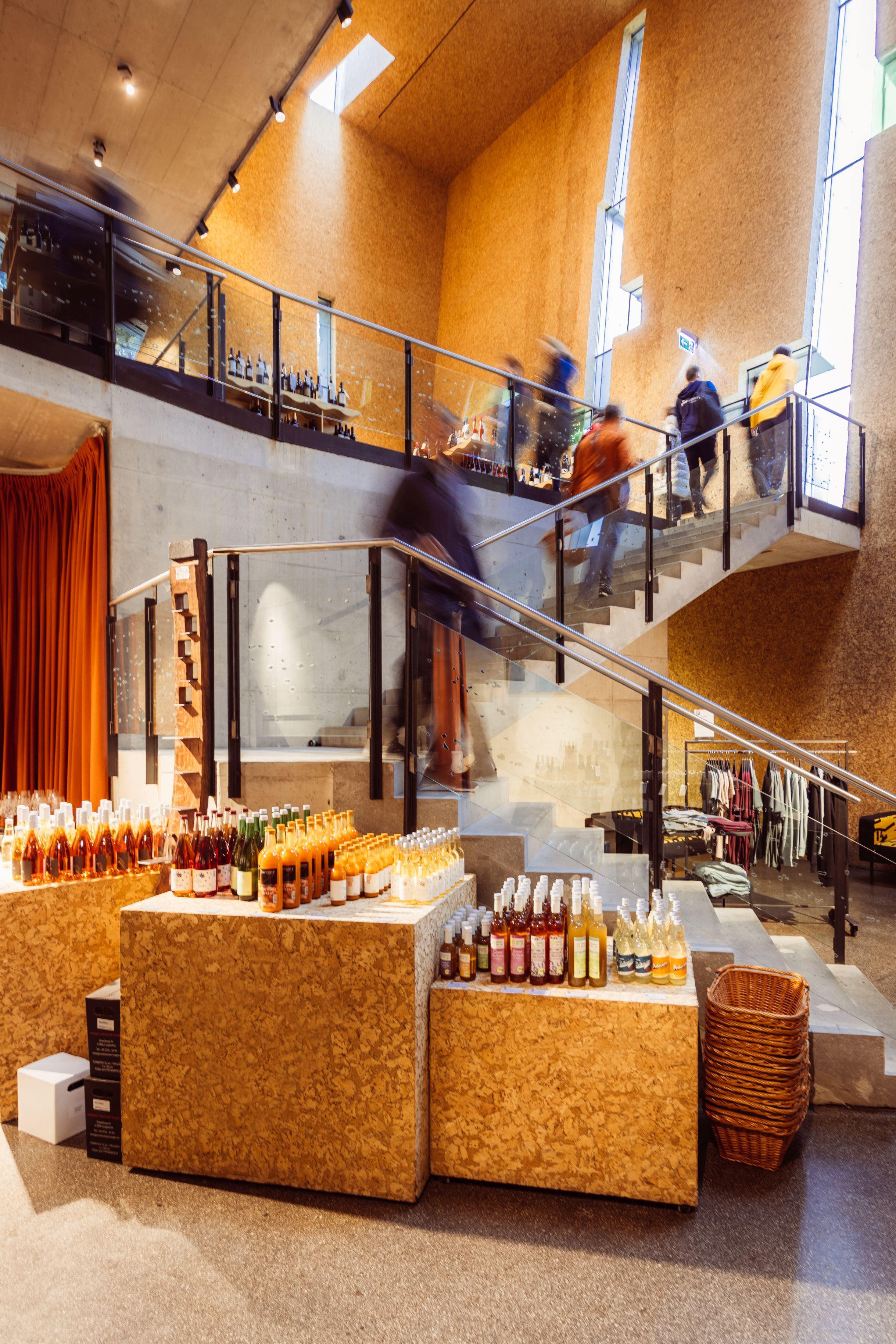 Interior view of Steininger Weinwelt, featuring a shop and wine bar: stairs, glass railing, wine bottles on display, and several people on the gallery.