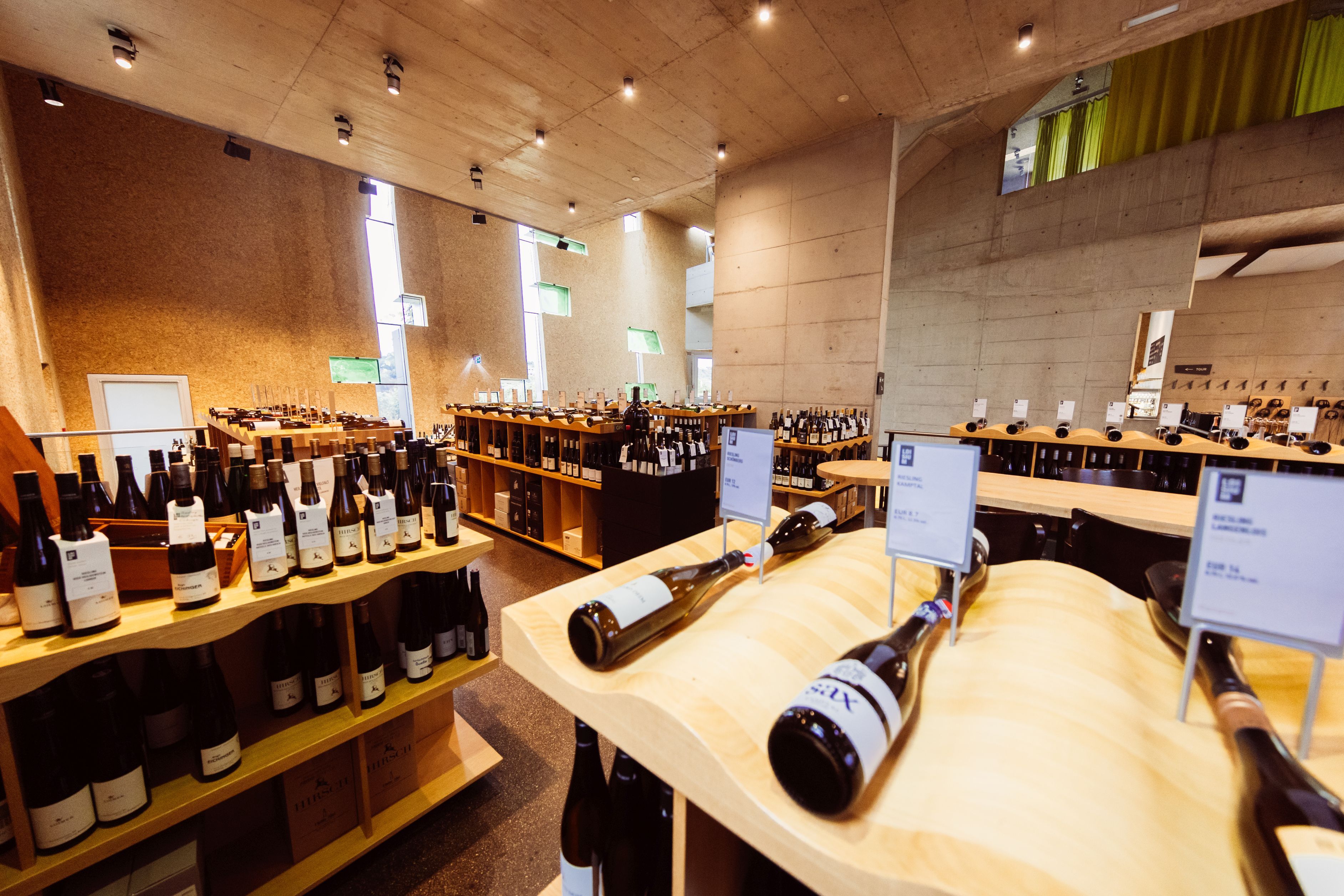 Interior view of the Steininger Weinwelt wine shop, featuring numerous bottles of wine on wooden shelves and tables in a modern concrete setting.