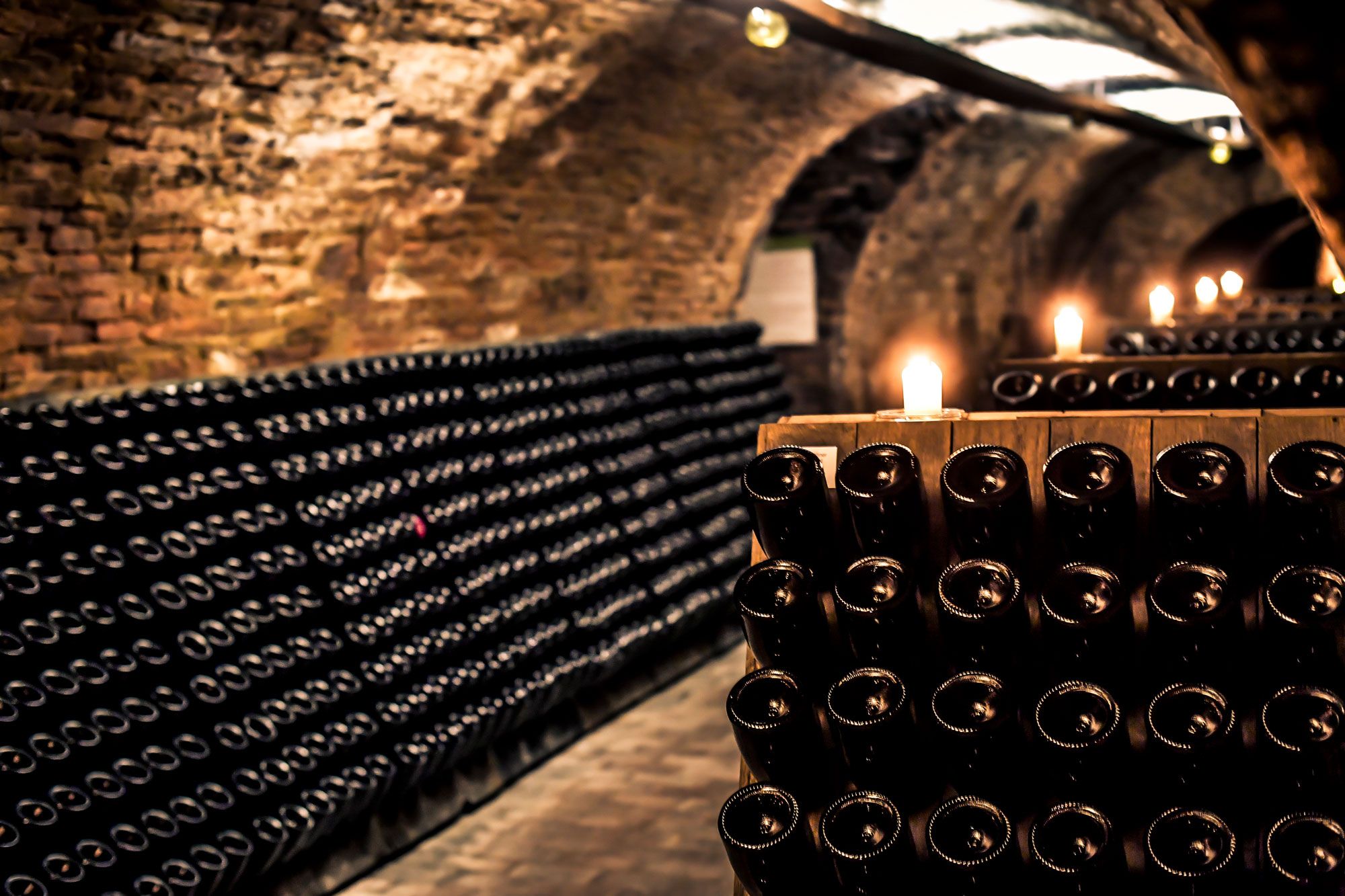 Weinregale mit Weinflaschen lagern waagerecht in einem schwach beleuchteten Backsteinkeller, wobei Kerzen im Vordergrund für warmes Licht sorgen.
