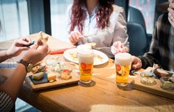 Ein Jausenbrett mit Käse und Aufstrichen im Hintergrund. Davor eine Person die Butter auf ihr Brot streicht. Daneben Bier von BrauSchneider. 