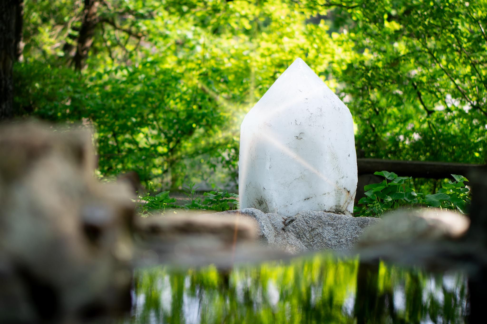 Ein großer weißer Kristall steht in einem grünen Wald, umgeben von Pflanzen und reflektiert in einem kleinen Teich.