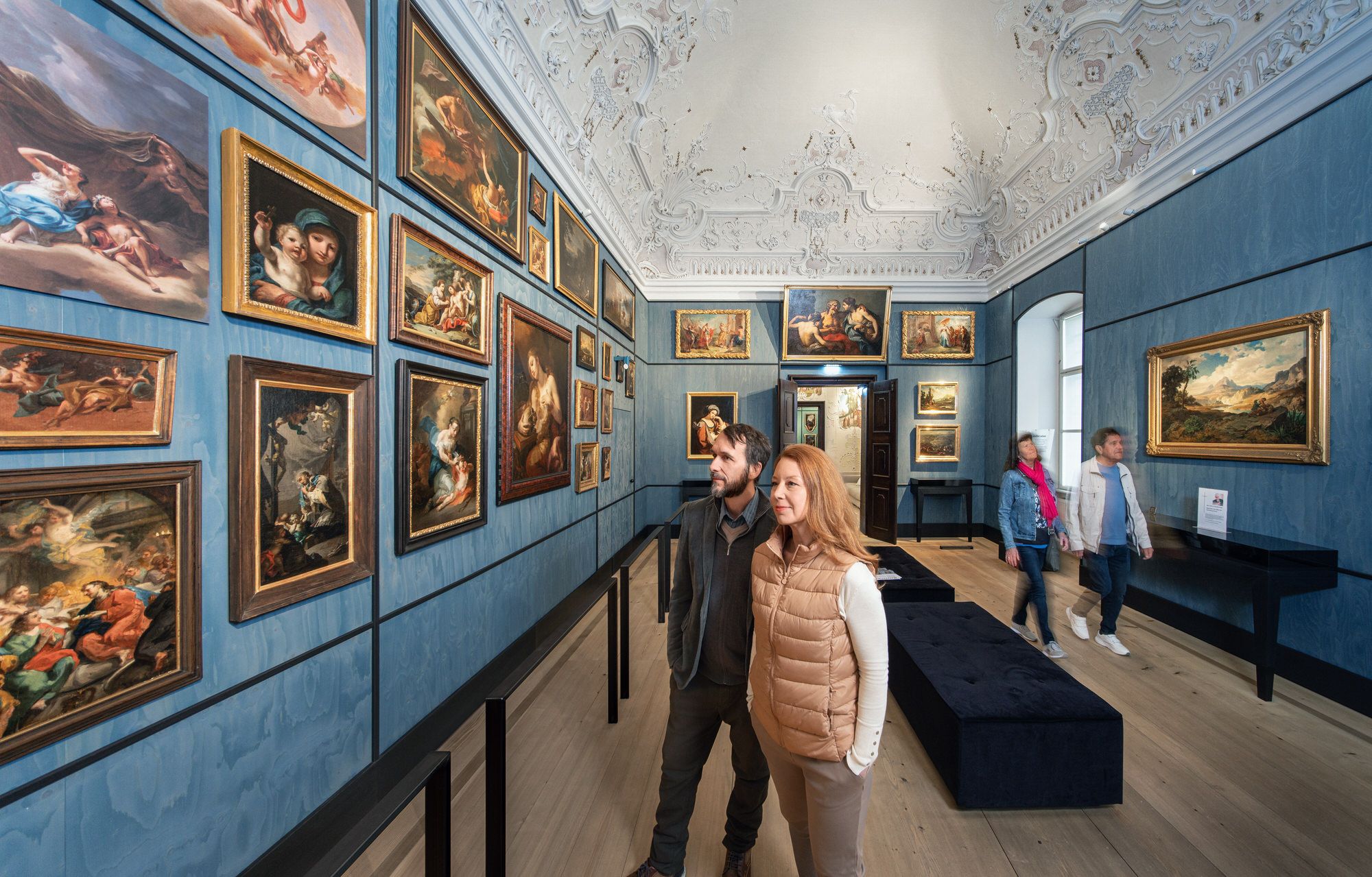 Besucher betrachten Gemälde in einem kunstvoll dekorierten Raum mit blauen Wänden und verziertem Deckenstuck.