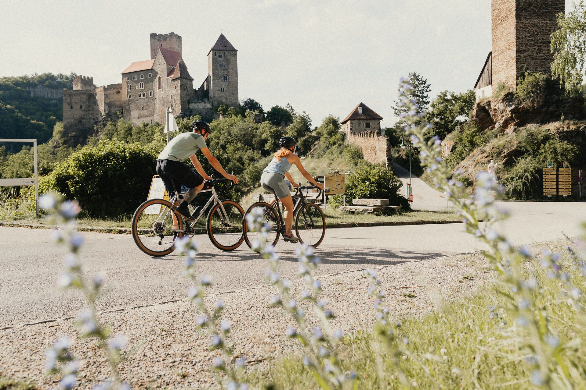 Zwei Radfahrer auf einer Straße vor der Burg Hardegg in einer grünen Landschaft.