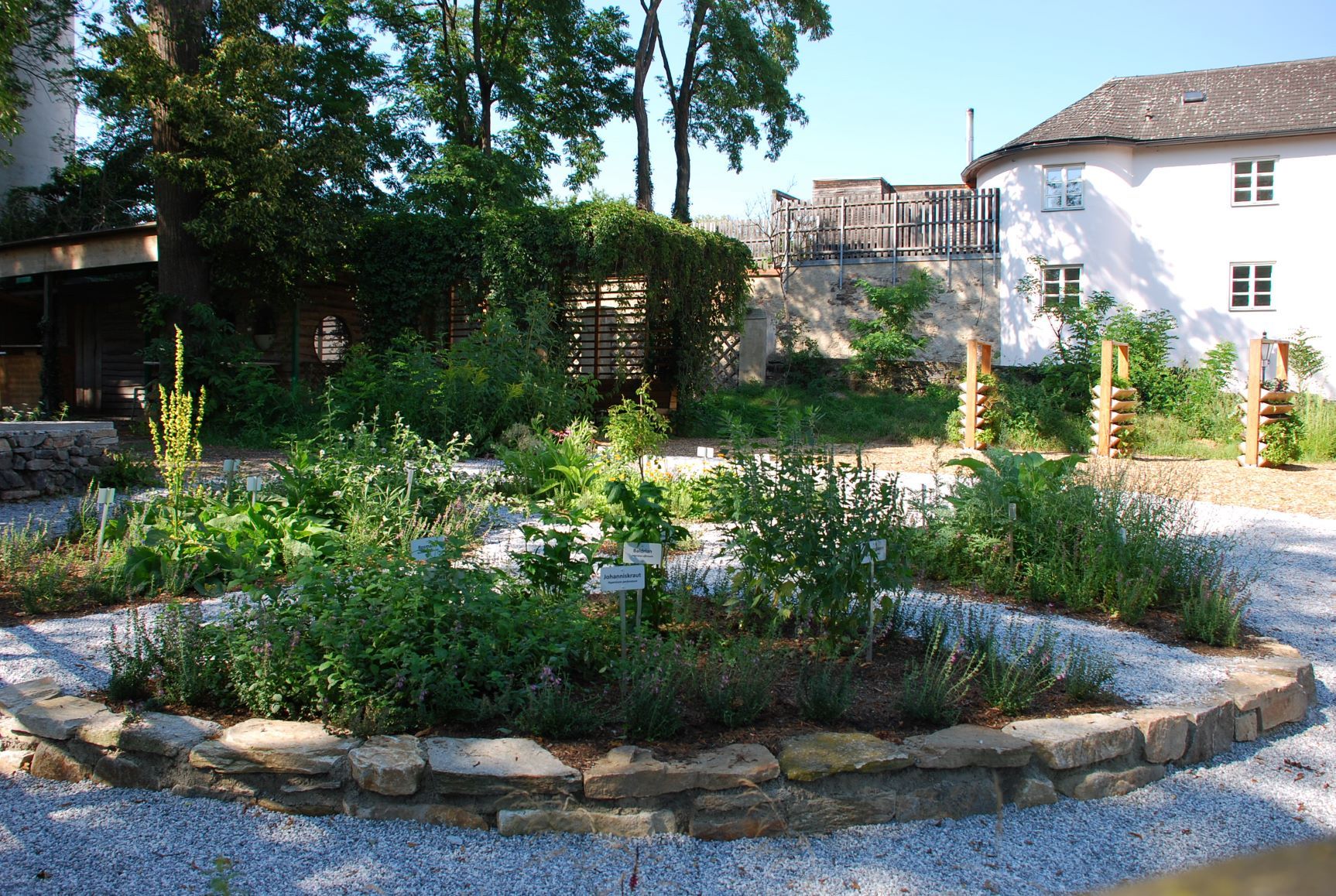 Ein gepflegter Garten mit verschiedenen Pflanzen und Schildern, umgeben von Bäumen und einem Gebäude im Hintergrund.