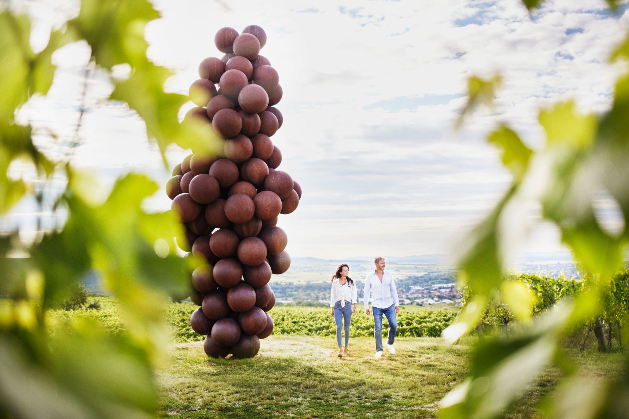 Zwei Personen spazieren neben einer großen Skulptur, der Riesenweintraube, am Weinweg in den Weinbergen.