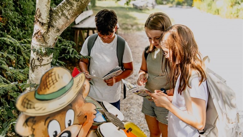Drei Kinder mit Rucksäcken lesen Karten neben einer Bärenfigur im Freien.