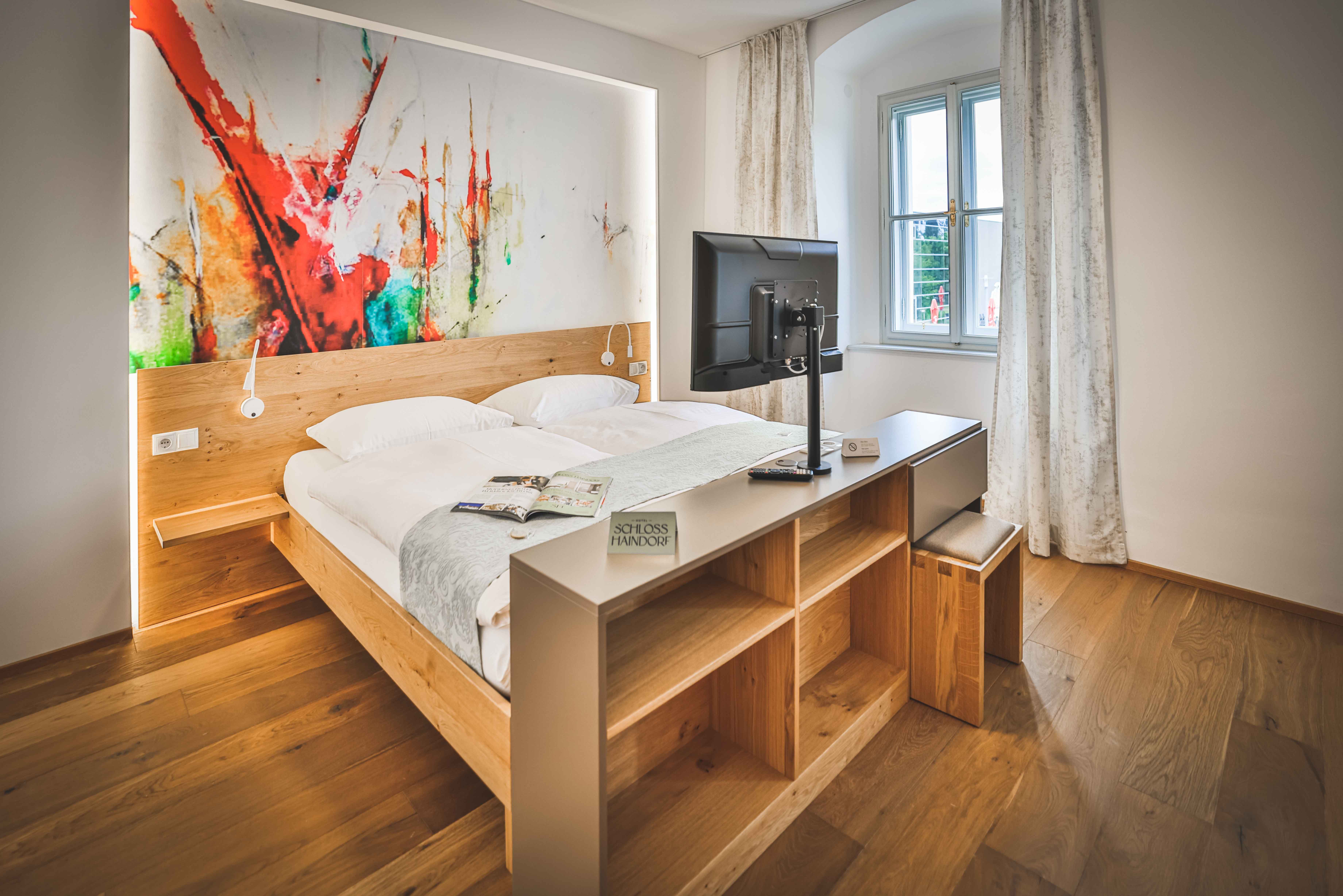 Modernes Hotelzimmer mit Doppelbett, abstraktem Kunstwerk an der Wand und einem Fernseher auf einem Regal.