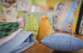 Zwei Stoffv&ouml;gel, einer gr&uuml;n und einer orange, auf einem Tisch mit Stoffen und N&auml;hutensilien.