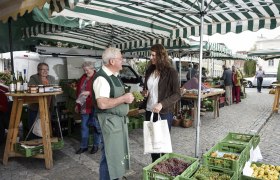 Menschen auf einem Wochenmarkt mit Obst- und Gemüseständen unter gestreiften Markisen.