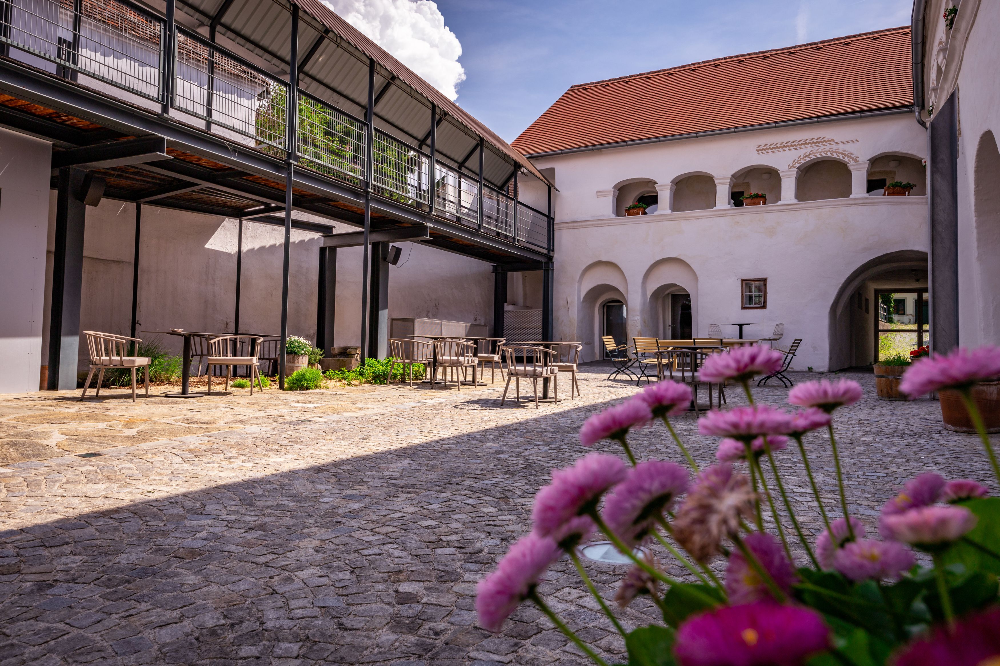 Ein Innenhof mit Kopfsteinpflaster, Tischen und Stühlen, umgeben von Gebäuden mit Arkaden und einem modernen Balkon.