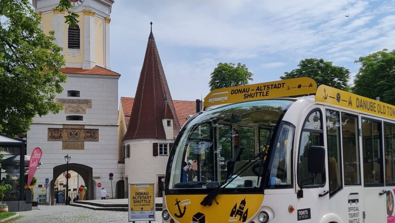 Ein wei&szlig;er Altstadt-Shuttlebus steht vor einem historischen Torbogen und einem Kirchturm.