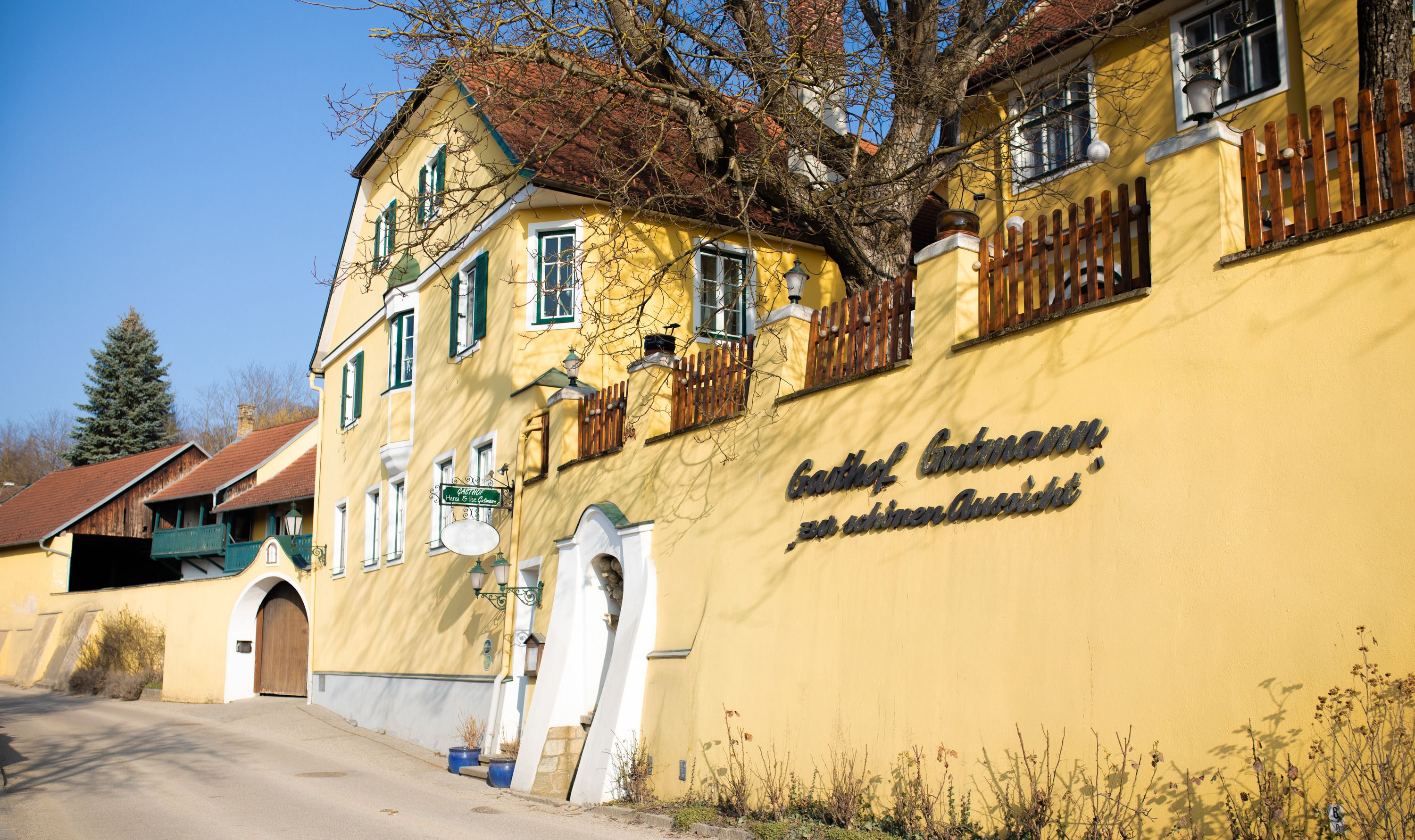 Gelbes Gasthaus mit grünen Fensterläden und Holzzaun, sonniger Tag.