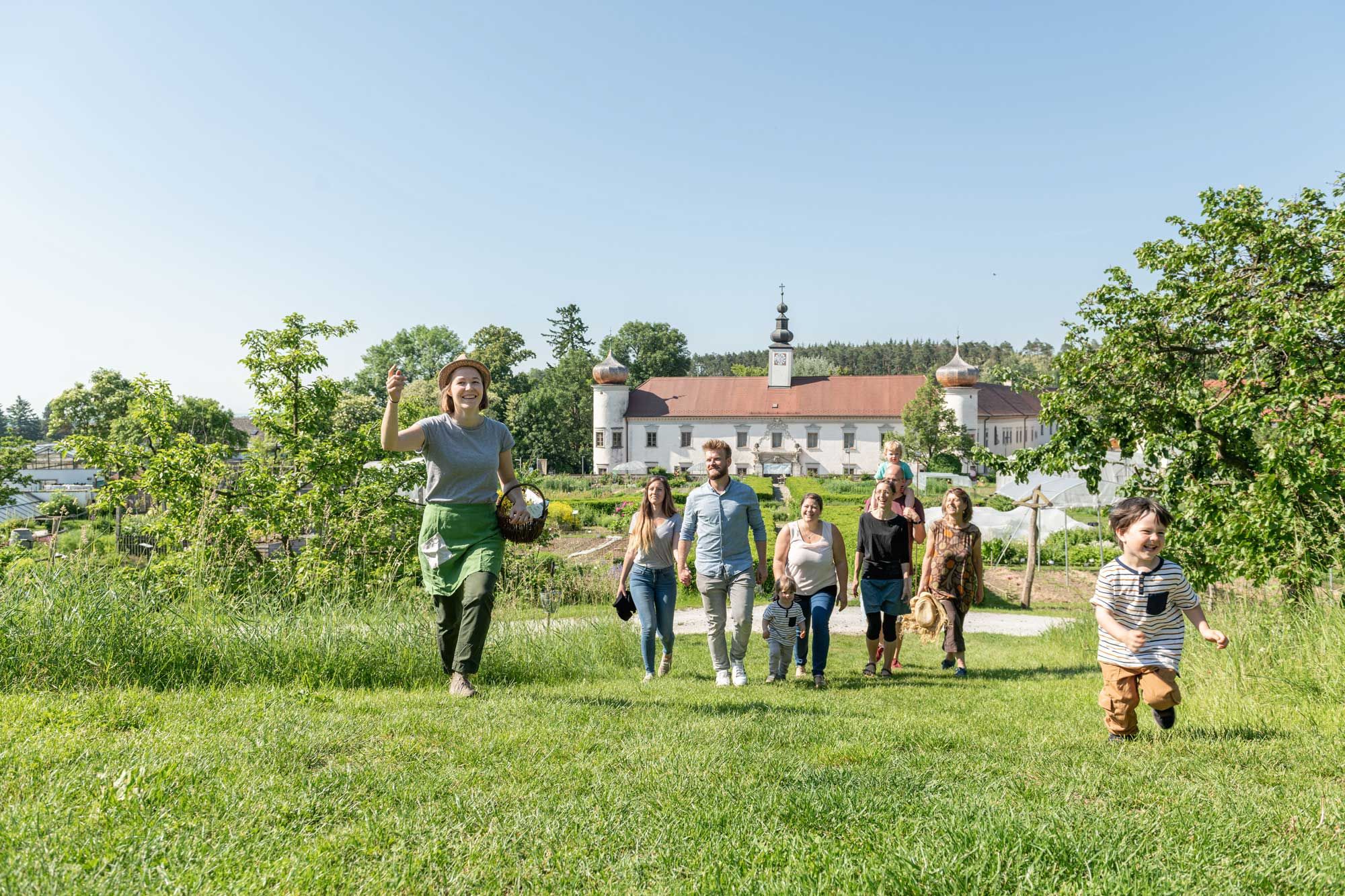 Eine Gruppe von Menschen geht über eine Wiese, im Hintergrund ein Gebäude mit Zwiebeltürmen.