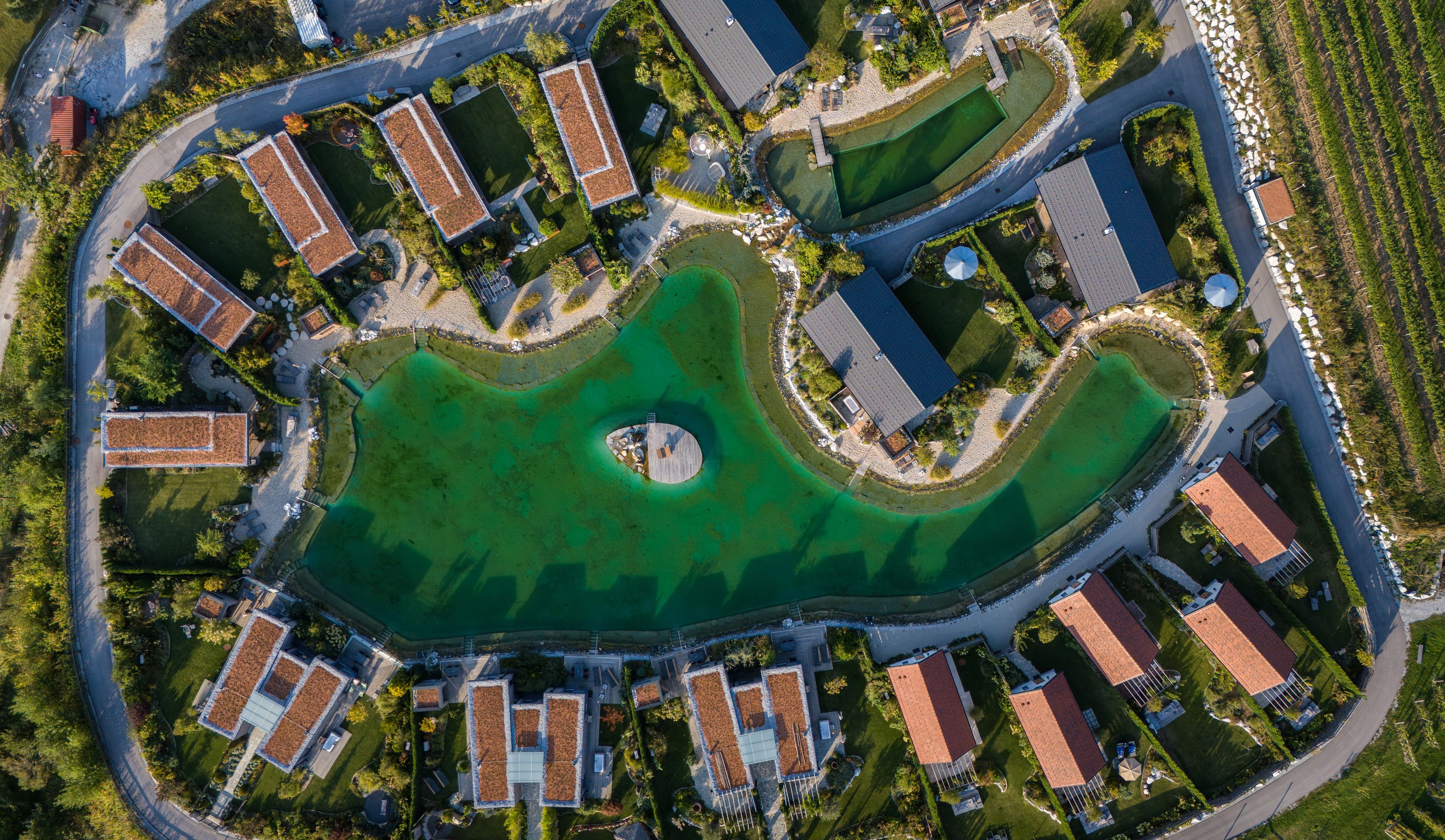 Luftaufnahme von Chalets um einen grünen See mit Steg in der Mitte.