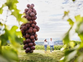 Zwei Personen spazieren an einer gro&szlig;en Skulptur aus kugelf&ouml;rmigen Elementen vorbei, umgeben von Weinbergen.