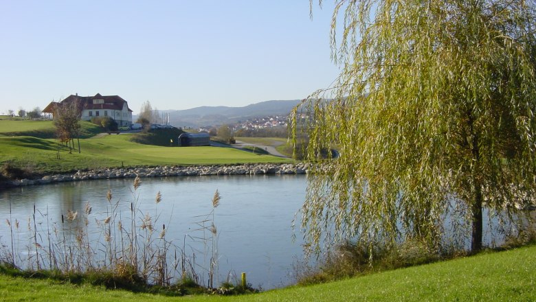 Landschaft mit Golfplatz, Teich und Geb&auml;ude im Hintergrund.