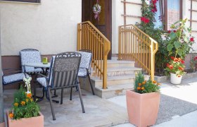 Eingangsbereich einer Ferienwohnung mit Terrasse, Tisch und St&uuml;hlen, Blumen und Gel&auml;nder.