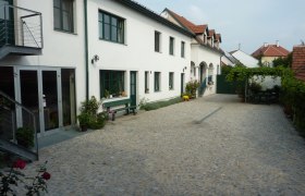 Innenhof eines Weinguts mit gepflastertem Boden, weißen Gebäuden und grünen Fensterrahmen.