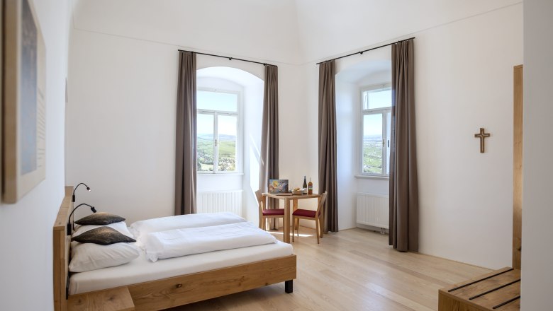 Helles Doppelzimmer mit Holzbett, Tisch und Stühlen, zwei Fenstern mit Vorhängen und einem Kreuz an der Wand.
