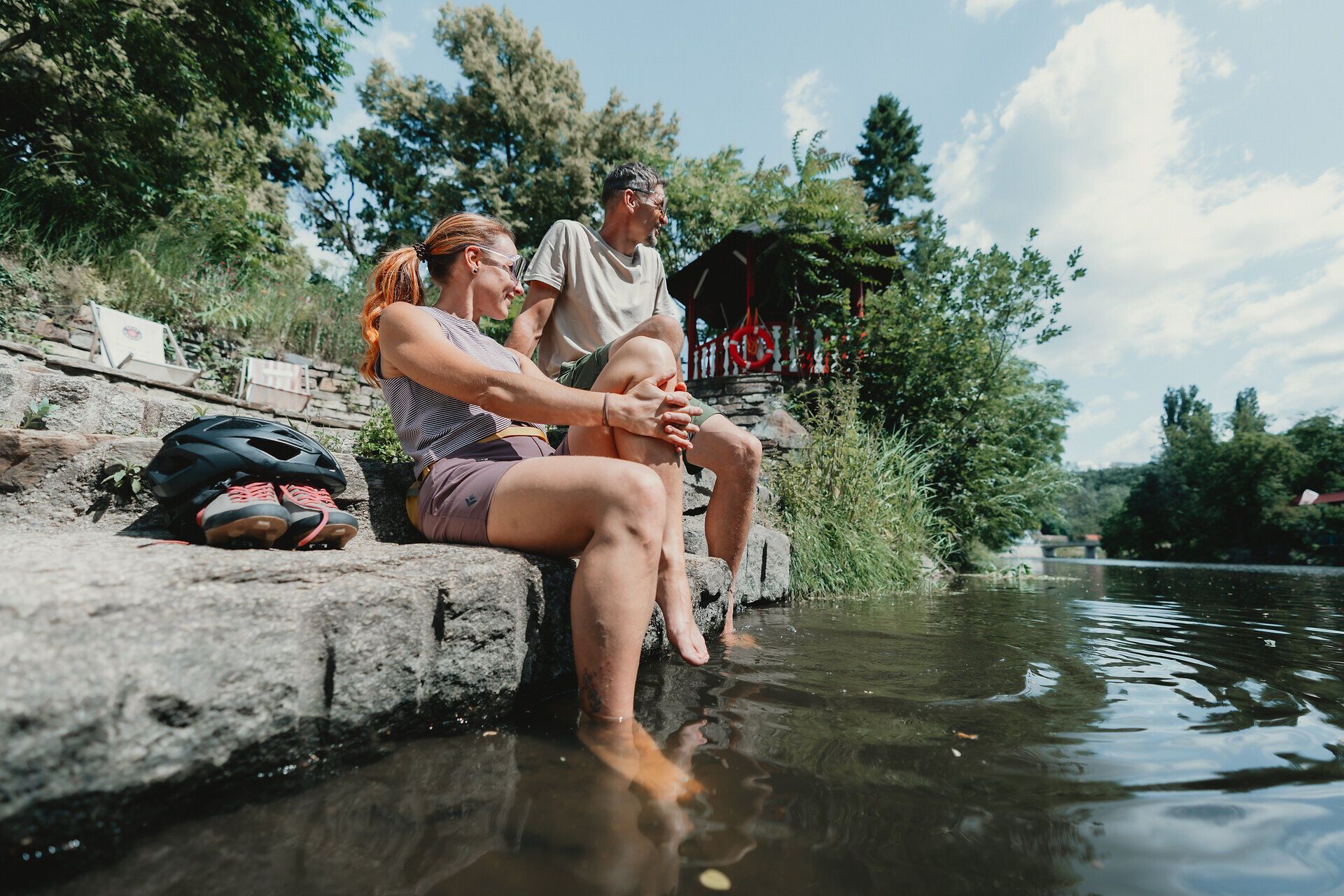 Zwei Personen sitzen auf einer Steintreppe am Flussufer und kühlen ihre Füße im Wasser. Ein Fahrradhelm und Schuhe liegen neben ihnen.