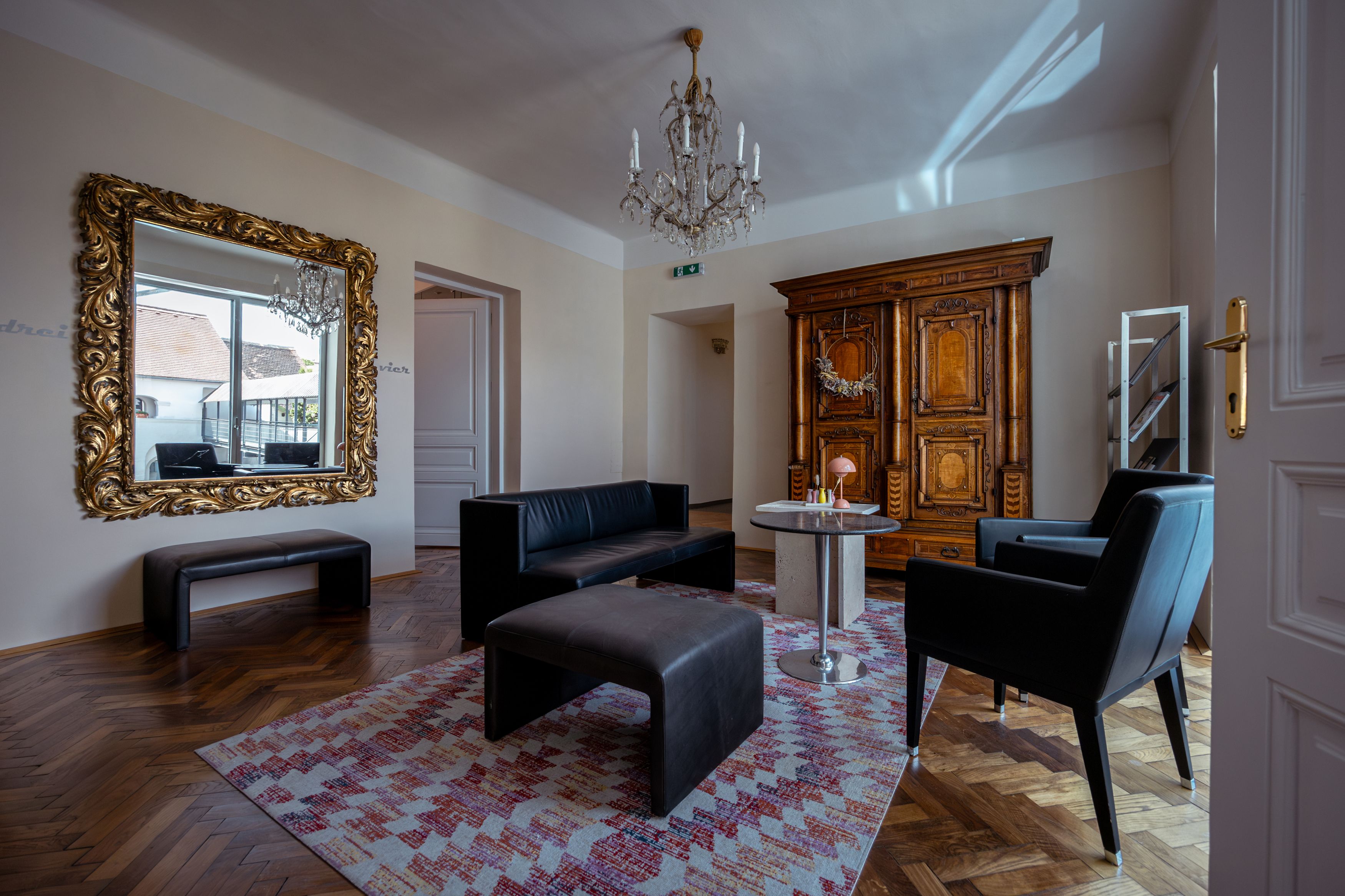Elegantes Wohnzimmer mit antikem Schrank, großem Spiegel, schwarzen Ledermöbeln und einem Kronleuchter.