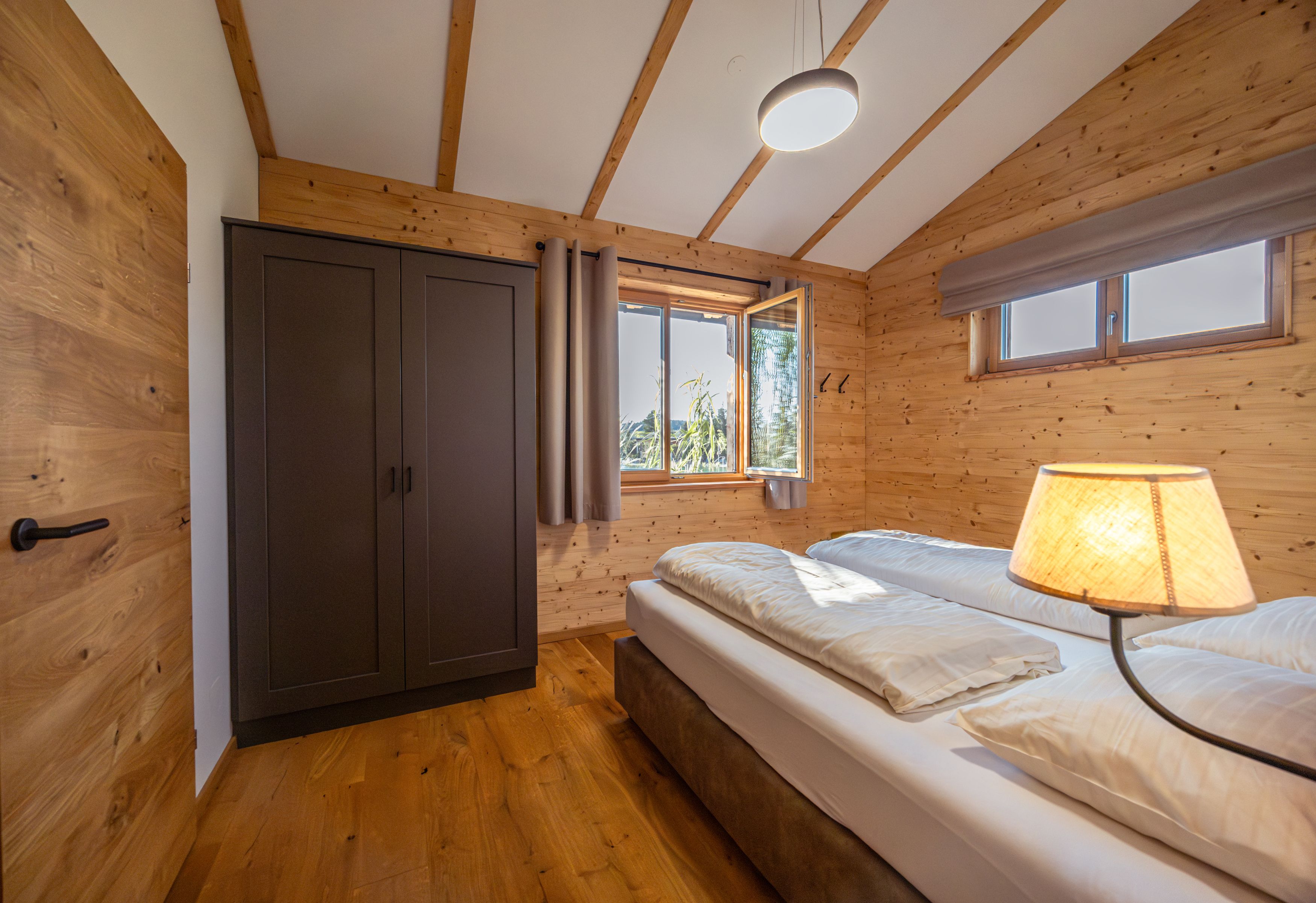 Innenansicht eines Schlafzimmers in einem Chalet mit Holzwänden, einem Doppelbett, einem Kleiderschrank und einem Fenster mit Blick nach draußen.