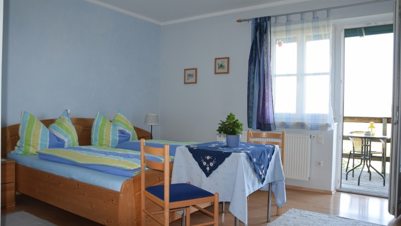 Gem&uuml;tliches G&auml;stezimmer mit Doppelbett, Tisch und St&uuml;hlen, blauen Vorh&auml;ngen und Zugang zu einem Balkon mit Sitzgelegenheit.