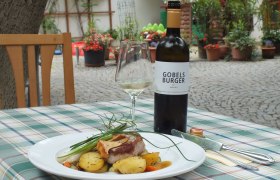 Ein Teller mit Fleisch und Kartoffeln auf einem Tisch im Freien, daneben eine Flasche Wein und ein Glas.