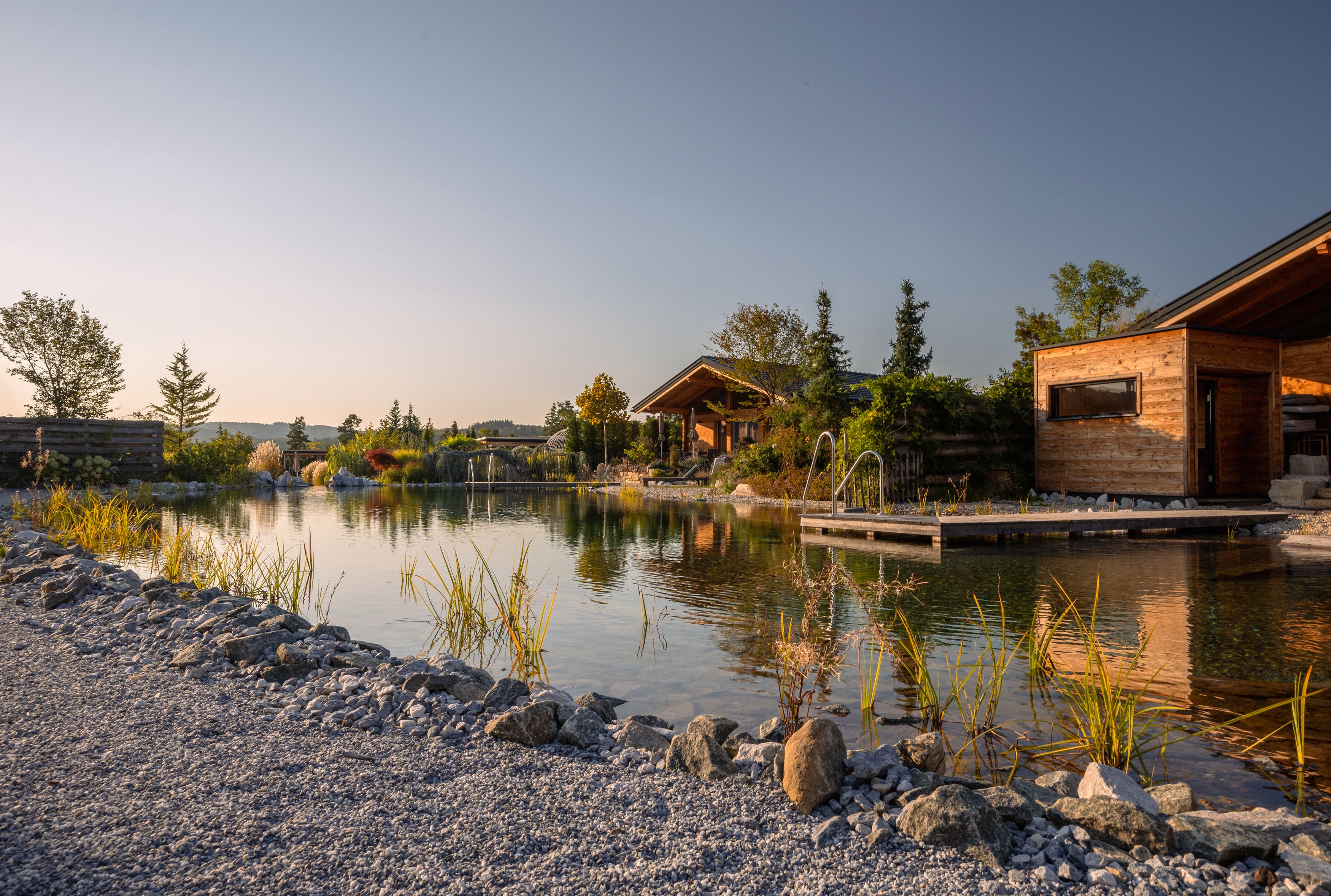 Holzchalets am Ufer eines kleinen Sees mit Steg und Pflanzen im Vordergrund.